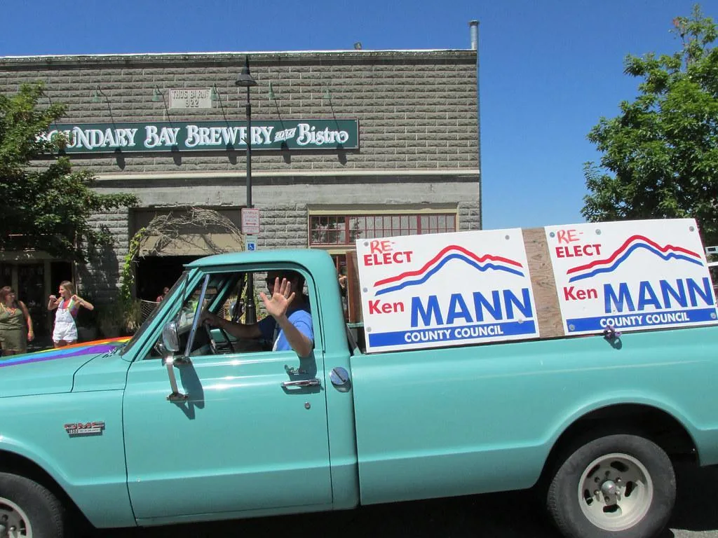 A turquise truck drives through town sporting political campaign signs.  The driver waves to the people on the street.